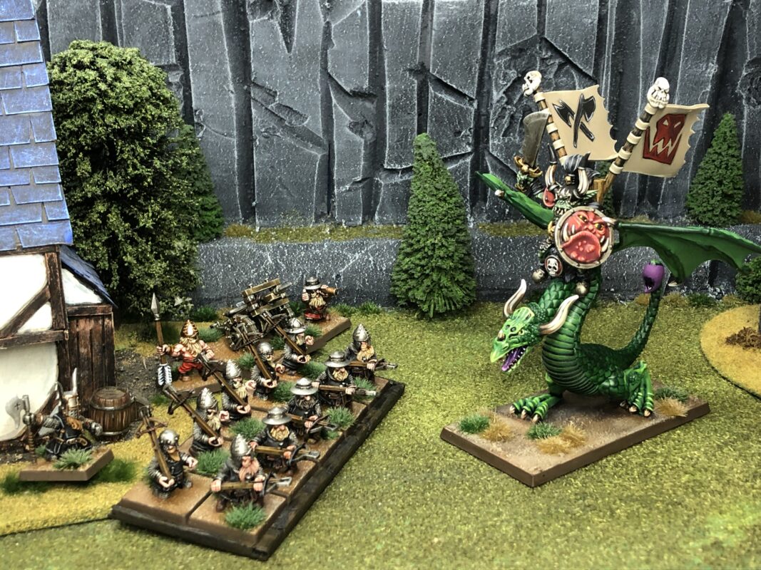 A fantasy warband facing a dragon outside a village